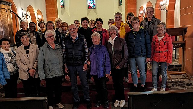 Gruppe von Menschen steht in einem Kirchenraum mit sichtbaren bunten Kirchenfenstern und roten Säulen.
