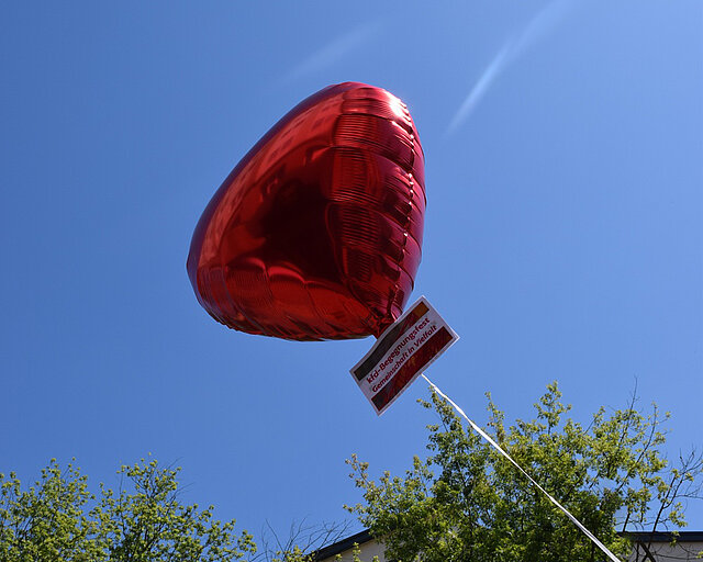 Roter herzförmiger Ballon vor blauem Himmel mit angehängtem Schild und Baumkronen im Hintergrund