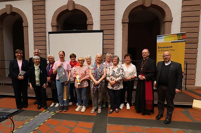 Gruppe von Menschen steht vor einer Leinwand und einem Banner mit der Aufschrift 'Ehrenamt im Bistum Trier' in einem Innenhof mit Steinbögen
