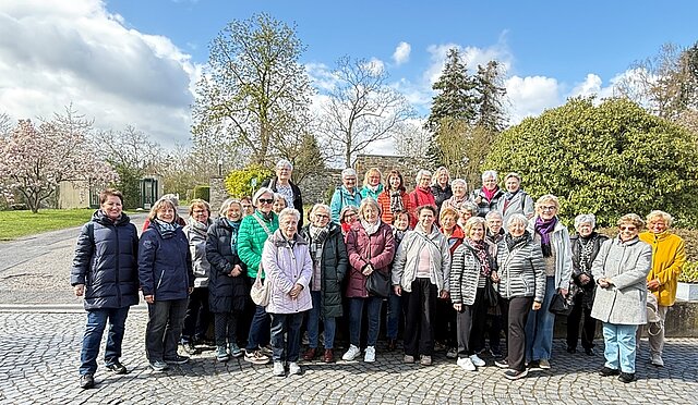 Eine Gruppe von Frauen in Jacken steht auf gepflastertem Platz vor Büschen und Bäumen bei klarem Himmel.